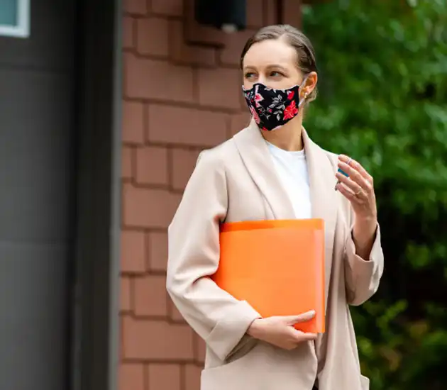 woman carrying an orange folder and wearing a mask standing outside a house