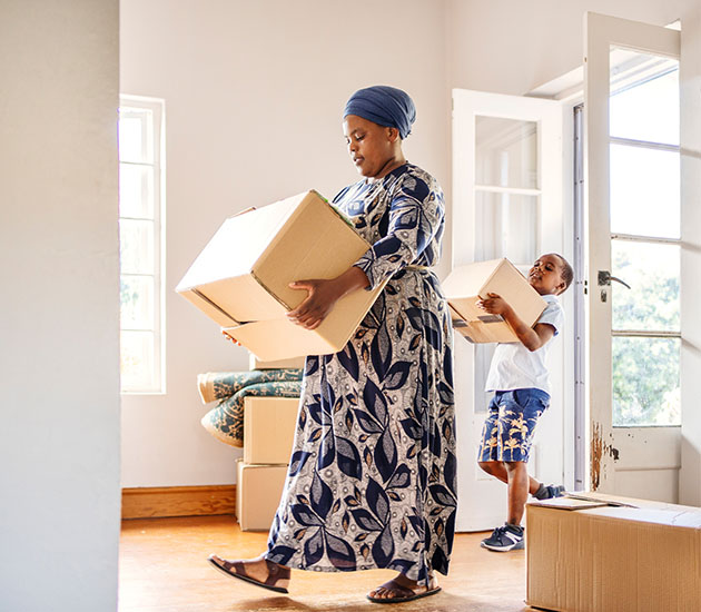 woman and her little son walking through a door into a living room carrying boxes while moving house